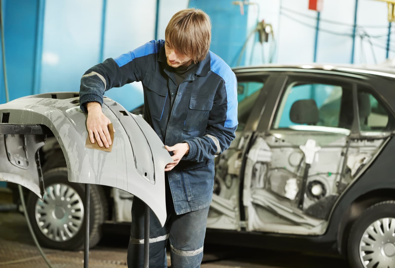 A body shop technician sands a primed bumper cover beside a stripped car in a repair bay.