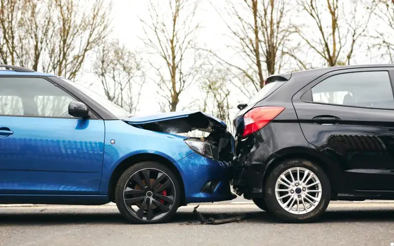 A blue hatchback with a crushed front end rear-ended a black hatchback on a road lined with bare trees, with broken plastic debris scattered on the pavement between them.