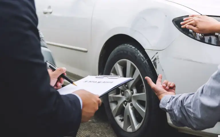 A person holds a clipboard labeled "insurance" with a pen while another gestures toward a scraped and dented front fender of a white car.